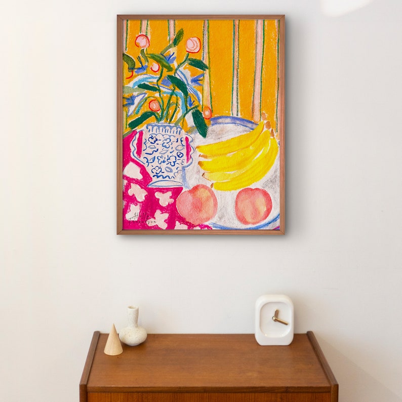 Op de afbeelding: Een ingelijst schilderij van een stilleven met een blauw-witte vaas gevuld met roze bloemen, een kom met gele bananen en twee roze perziken op een roze-witte tafelkleed met een geel-wit gestreepte achtergrond.