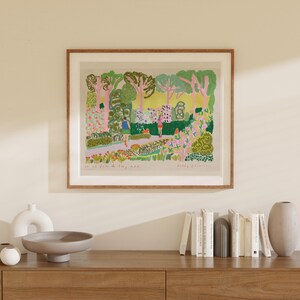 May include: A framed art print depicting a colorful garden scene with figures. The artwork features a variety of trees, flowers, and foliage in shades of green, pink, and yellow. The print is displayed in a wooden frame and is mounted on a light-colored wall.