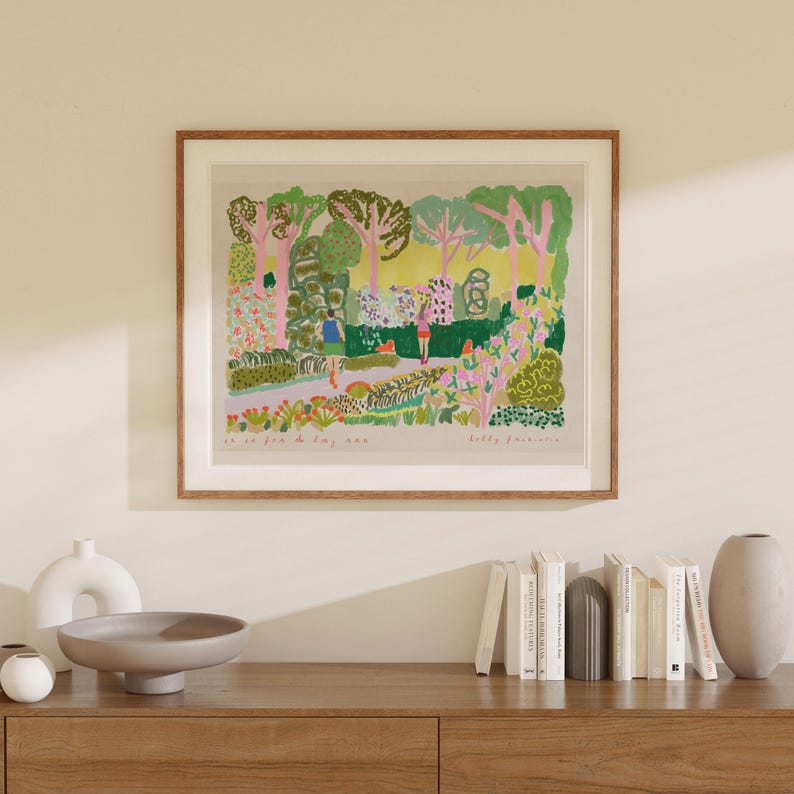May include: A framed art print depicting a colorful garden scene with figures. The artwork features a variety of trees, flowers, and foliage in shades of green, pink, and yellow. The print is displayed in a wooden frame and is mounted on a light-colored wall.