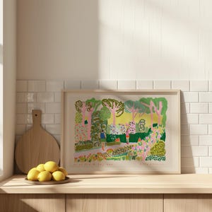 May include: A framed art print featuring a colorful garden scene with figures and trees. The artwork has a light wood frame and is displayed on a wooden surface. The print includes text at the bottom and is set against a white tile background.
