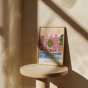 K&ouml;nnte beinhalten: Eine farbenfrohe Illustration von Amsterdam, Niederlande, mit einem Farbschema in Rosa, Gelb und Blau. Die Illustration zeigt einen Kanal mit Booten und Geb&auml;uden am Rande. Der Text "AMSTERDAM" befindet sich am unteren Rand der Illustration.