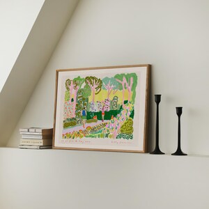 May include: A framed art print with a colorful garden scene, featuring trees, flowers, and figures. The print is in a light wood frame, with two black candle holders to the right. A stack of books is on the left.