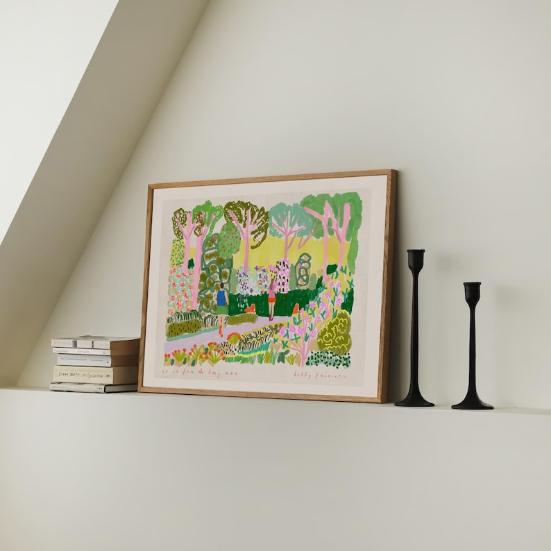 May include: A framed art print with a colorful garden scene, featuring trees, flowers, and figures. The print is in a light wood frame, with two black candle holders to the right. A stack of books is on the left.