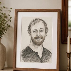Puede incluir: Un retrato a lápiz enmarcado de una persona con el pelo corto y barba, vistiendo una camiseta tipo henley oscura. El dibujo está realizado en grafito sobre papel blanco roto, dentro de un marco de madera sencillo con un paspartú blanco.