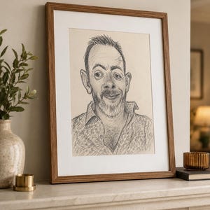 Puede incluir: Un retrato a lápiz enmarcado de un hombre con barba y camisa estampada. La obra de arte está en un marco de madera con un passe-partout blanco. El retrato se exhibe en una repisa blanca, con un jarrón y objetos decorativos en el fondo.