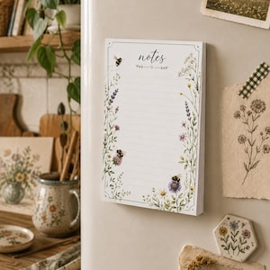 Puede incluir: Un bloc de notas rectangular con la palabra notes impresa en la parte superior, que presenta líneas horizontales para escribir. El borde está decorado con ilustraciones de flores silvestres, follaje verde y tres abejorros en tonos amarillo, morado y blanco.