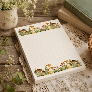 May include: A rectangular notepad with horizontal lines for writing, featuring a watercolor illustration of various brown and orange mushrooms surrounded by green foliage printed along the top and bottom edges on a white paper background.