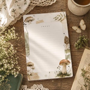 Può includere: Un blocco note rettangolare con linee orizzontali e la parola NOTES stampata in alto. La carta presenta un'illustrazione botanica con vari funghi, fogliame verde e piccoli punti neri su uno sfondo bianco con accenti color beige.