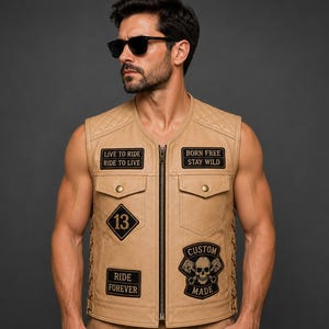 May include: A tan leather vest featuring a quilted shoulder design, a central zipper, and multiple black embroidered patches. The patches display text including "LIVE TO RIDE," "BORN FREE," "13," "RIDE FOREVER," and "CUSTOM MADE" with a skull graphic.