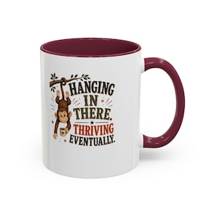 May include: A white ceramic mug with a maroon interior and handle. The exterior features a brown monkey hanging from a branch, holding a small mug with a heart. The text reads: HANGING IN THERE. THRIVING EVENTUALLY. The design includes small stars.