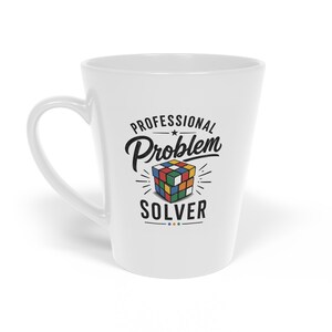 May include: A white ceramic latte mug featuring a graphic of a colourful multi-coloured puzzle cube. The text "PROFESSIONAL Problem SOLVER" is printed in black above and below the cube illustration, with small decorative lines surrounding the design.