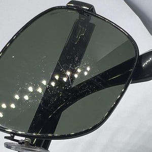 May include: A pair of sunglasses with dark, reflective lenses and a black frame. The lenses have a visible surface texture with light reflections and fine scratches. The frame features a thin, dark metal construction with clear nose pads.