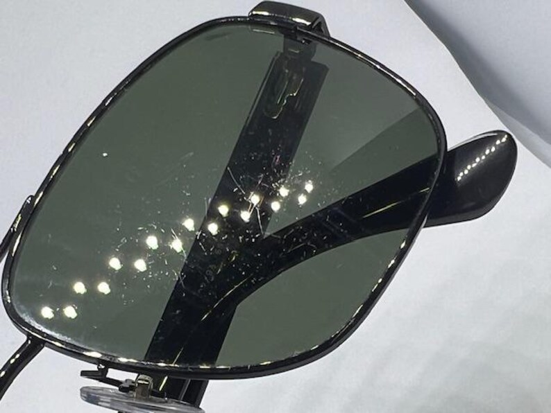 May include: A pair of sunglasses with dark, reflective lenses and a black frame. The lenses have a visible surface texture with light reflections and fine scratches. The frame features a thin, dark metal construction with clear nose pads.