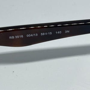 Pode incluir: Uma haste de &oacute;culos de sol em tom castanho escuro transl&uacute;cido com texto impresso a branco no lado interior. O texto diz RB 3516 004/13 59 15 140 3N, indicando o n&uacute;mero do modelo, o c&oacute;digo da cor e as medidas da arma&ccedil;&atilde;o em mil&iacute;metros.