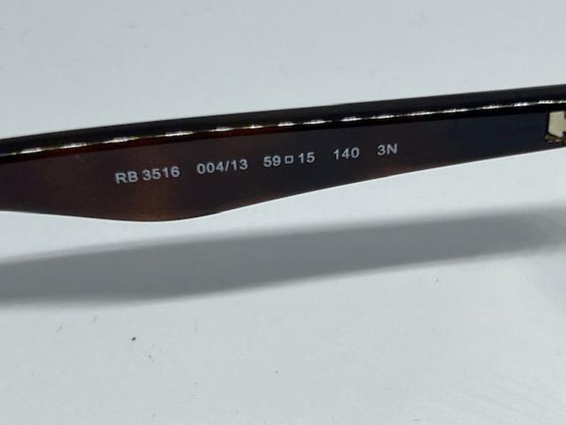 Pode incluir: Uma haste de &oacute;culos de sol em tom castanho escuro transl&uacute;cido com texto impresso a branco no lado interior. O texto diz RB 3516 004/13 59 15 140 3N, indicando o n&uacute;mero do modelo, o c&oacute;digo da cor e as medidas da arma&ccedil;&atilde;o em mil&iacute;metros.