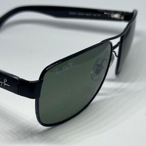 May include: Black Ray-Ban sunglasses featuring a rectangular frame design with dark green polarized lenses. The Ray-Ban logo is printed in white on the upper corner of the right lens and on the black temples. The frame includes a double bridge.