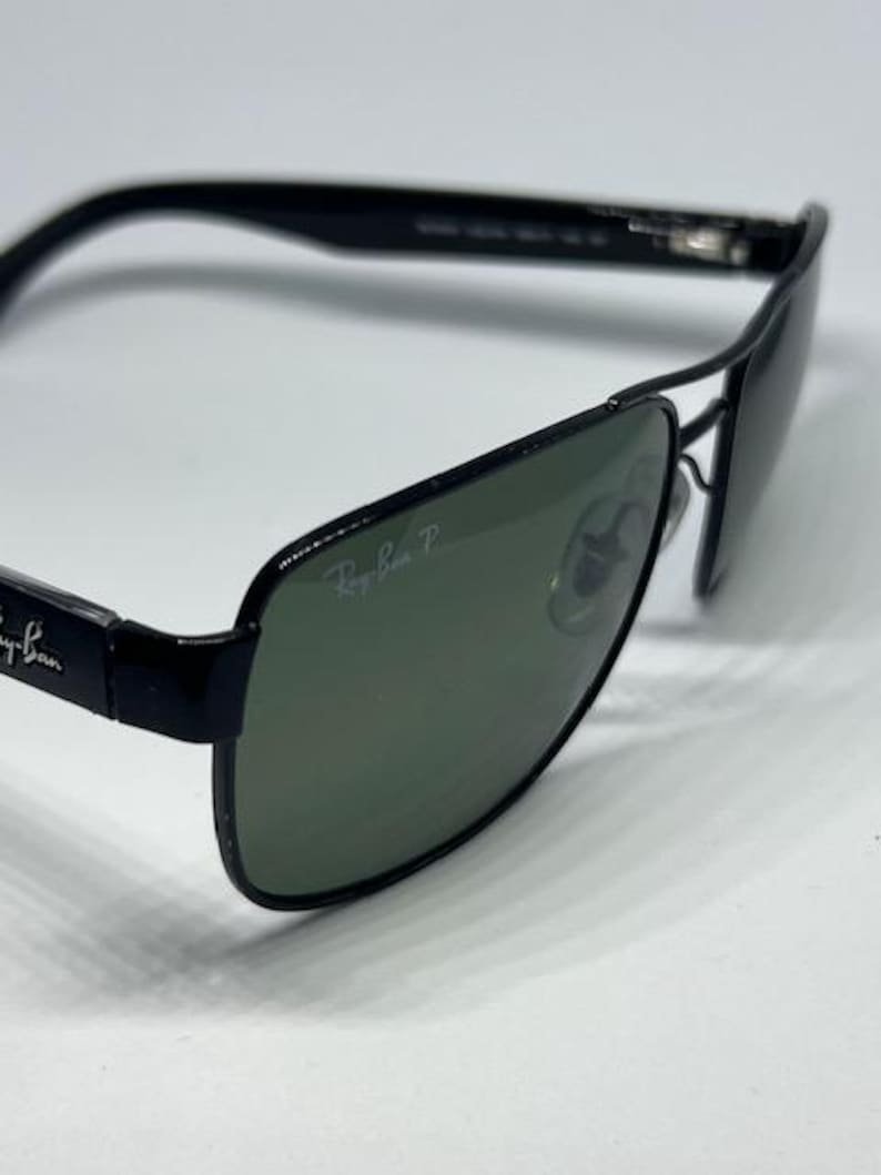 May include: Black Ray-Ban sunglasses featuring a rectangular frame design with dark green polarized lenses. The Ray-Ban logo is printed in white on the upper corner of the right lens and on the black temples. The frame includes a double bridge.