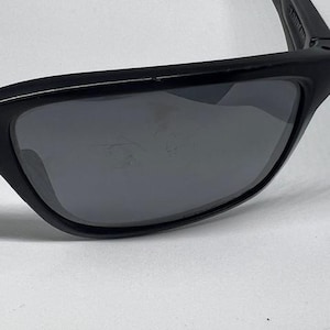 May include: Black sunglasses featuring a rectangular frame and dark gray lenses. The frame has a smooth matte finish, and the lenses show visible surface scratches. The word 'JANTZEN' is printed in white on the inner side of the temple arm.
