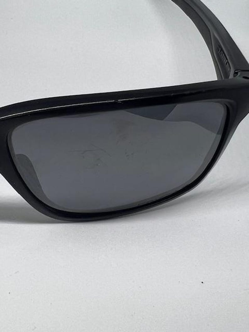 May include: Black sunglasses featuring a rectangular frame and dark gray lenses. The frame has a smooth matte finish, and the lenses show visible surface scratches. The word 'JANTZEN' is printed in white on the inner side of the temple arm.