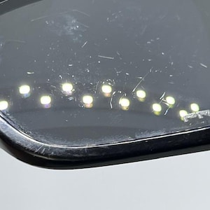 May include: A pair of black sunglasses with dark, scratched lenses. The lenses feature a series of small, bright light reflections arranged in a curved line across the surface. The word PRIZM is etched into the lower corner of the right lens.