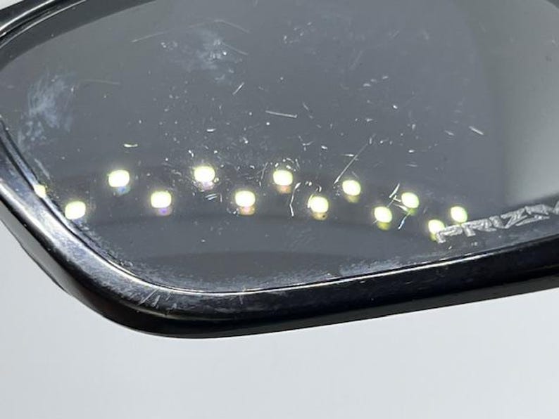 May include: A pair of black sunglasses with dark, scratched lenses. The lenses feature a series of small, bright light reflections arranged in a curved line across the surface. The word PRIZM is etched into the lower corner of the right lens.