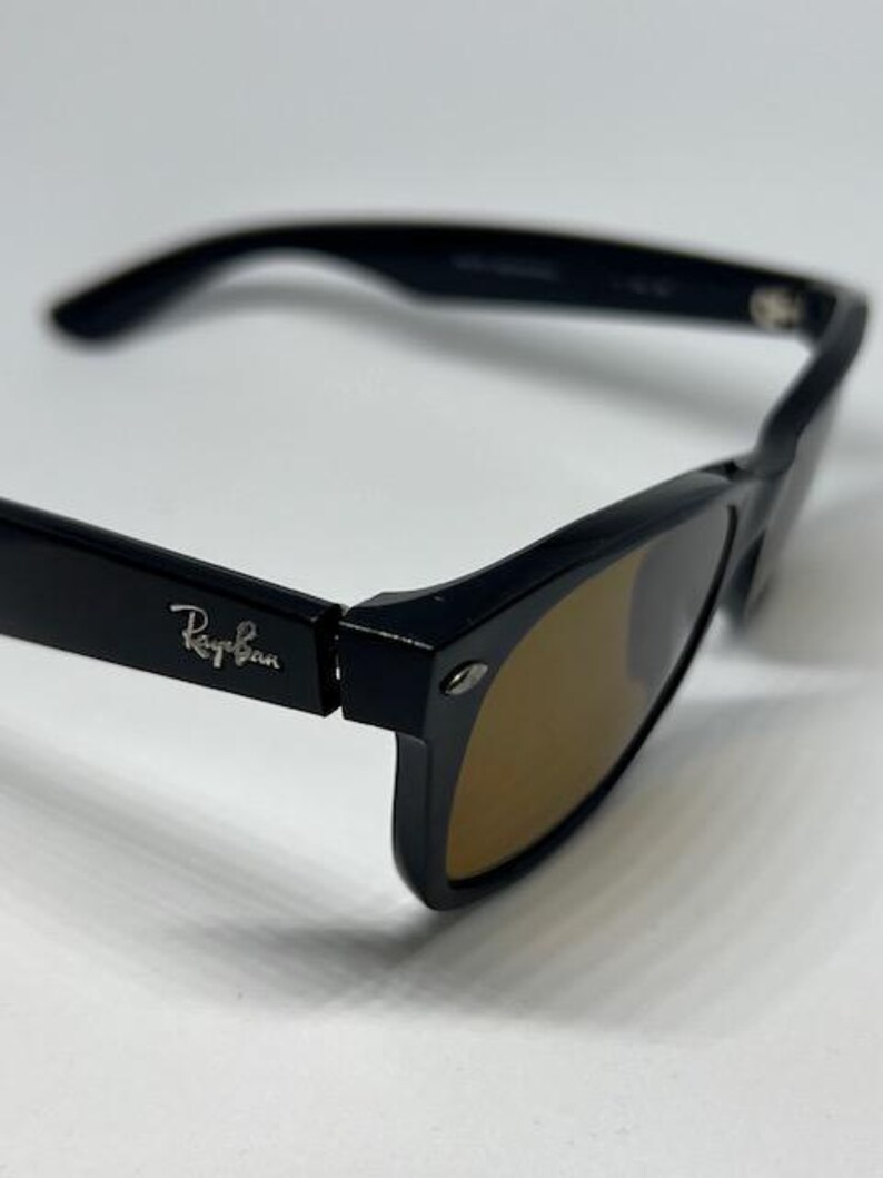 Puede incluir: Gafas de sol negras con montura gruesa y lentes tintadas en marr&oacute;n. La patilla presenta el logotipo plateado de Ray-Ban en relieve en el lateral. El dise&ntilde;o incluye una forma rectangular cl&aacute;sica con un detalle de bisagra met&aacute;lica.