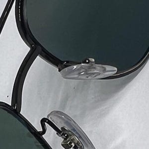 May include: A pair of black metal frame sunglasses featuring dark tinted lenses and clear silicone nose pads. The frame has a thin, angular design with a double bridge structure visible at the top. The nose pads are attached to thin metal arms.