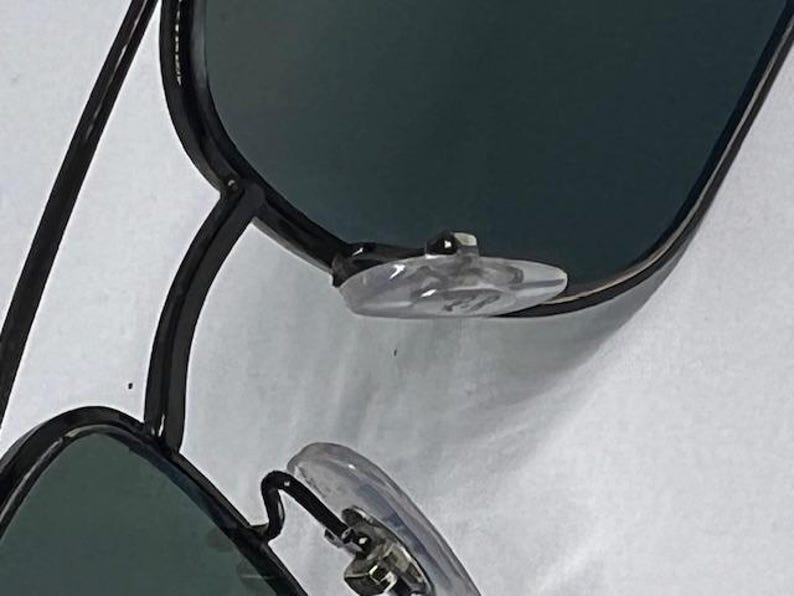 May include: A pair of black metal frame sunglasses featuring dark tinted lenses and clear silicone nose pads. The frame has a thin, angular design with a double bridge structure visible at the top. The nose pads are attached to thin metal arms.