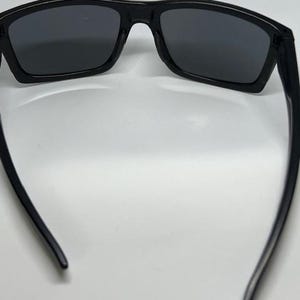 May include: A pair of black sunglasses with dark, rectangular lenses and a smooth, solid black frame. The temples are curved inward, and the bridge connects the two lenses in a continuous, streamlined design, providing a clear view of the interior.