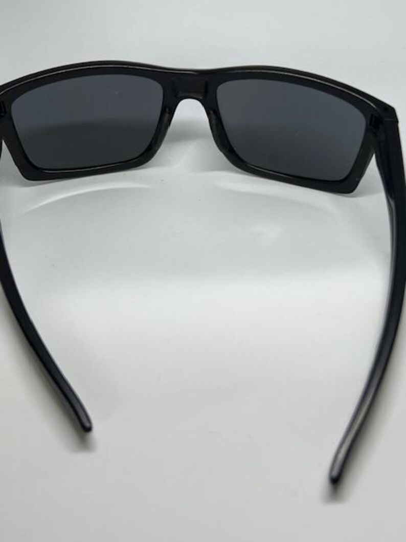 May include: A pair of black sunglasses with dark, rectangular lenses and a smooth, solid black frame. The temples are curved inward, and the bridge connects the two lenses in a continuous, streamlined design, providing a clear view of the interior.