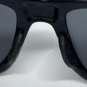 May include: A close-up view of the bridge and nose pads of black sunglasses. The frame is made of a glossy black material, and the nose pads are constructed from a textured, matte black rubberized material designed for grip and comfort.