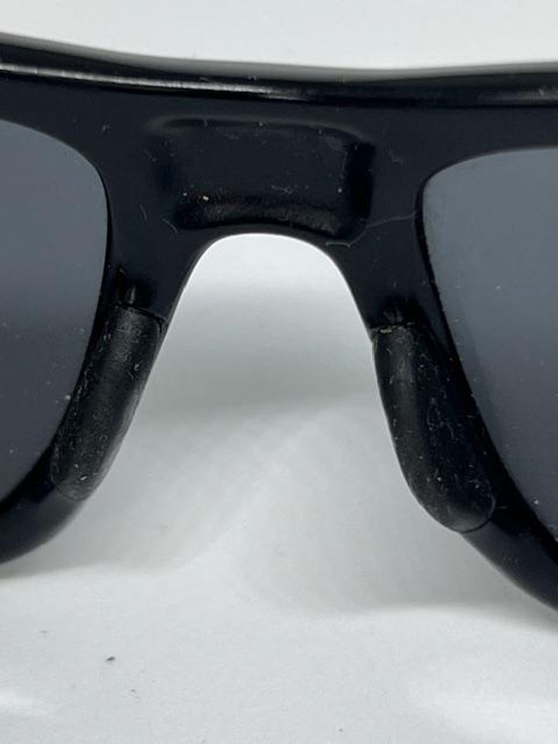 May include: A close-up view of the bridge and nose pads of black sunglasses. The frame is made of a glossy black material, and the nose pads are constructed from a textured, matte black rubberized material designed for grip and comfort.