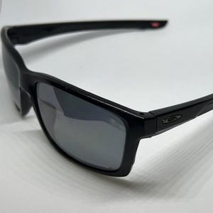 May include: Black sunglasses featuring a rectangular frame and dark, reflective lenses. The temples are sleek and black, displaying a small, metallic oval logo on the side near the hinge. The overall design is streamlined with a glossy finish.