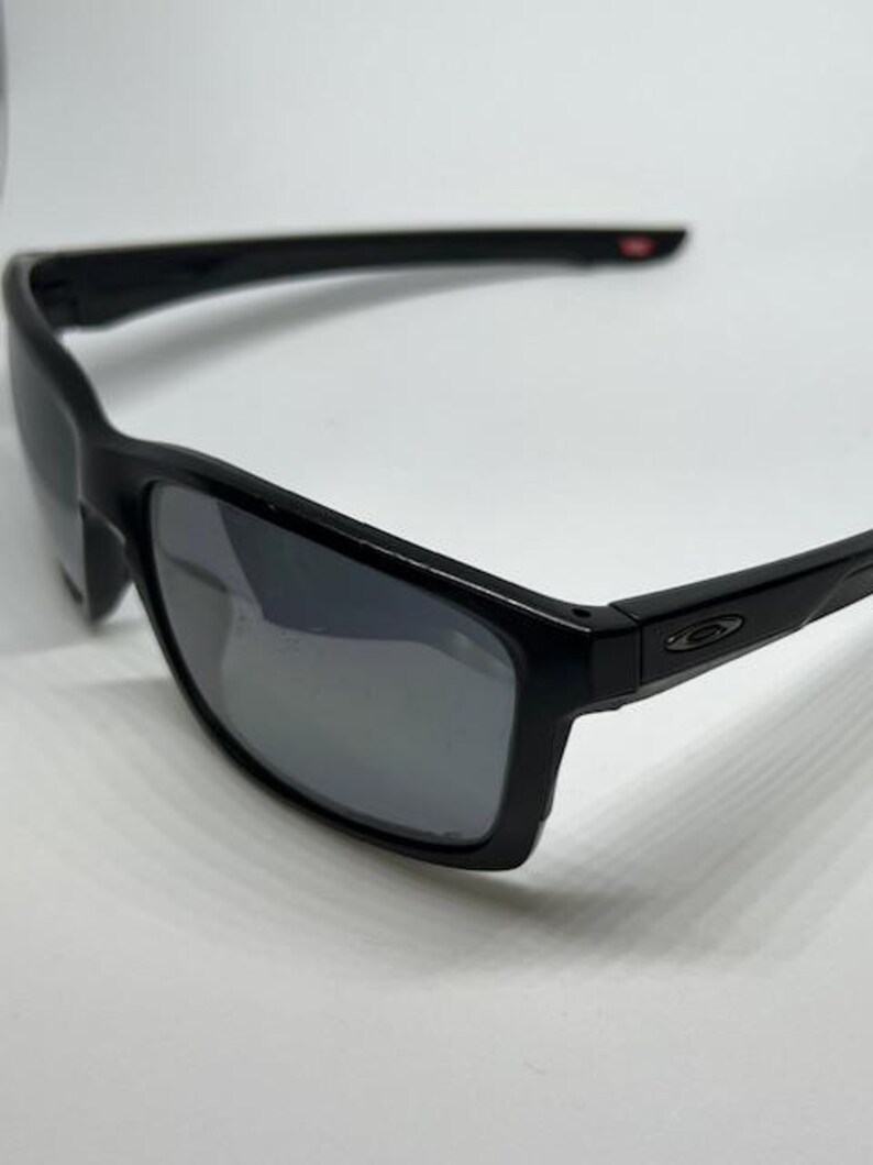 May include: Black sunglasses featuring a rectangular frame and dark, reflective lenses. The temples are sleek and black, displaying a small, metallic oval logo on the side near the hinge. The overall design is streamlined with a glossy finish.