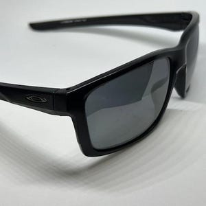 May include: Black sunglasses featuring a rectangular frame design with dark, reflective lenses. The temples display a small, metallic Oakley logo emblem on the side, and the overall construction consists of a smooth, solid black plastic material.