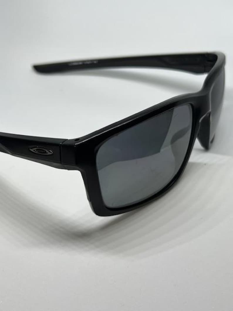 May include: Black sunglasses featuring a rectangular frame design with dark, reflective lenses. The temples display a small, metallic Oakley logo emblem on the side, and the overall construction consists of a smooth, solid black plastic material.