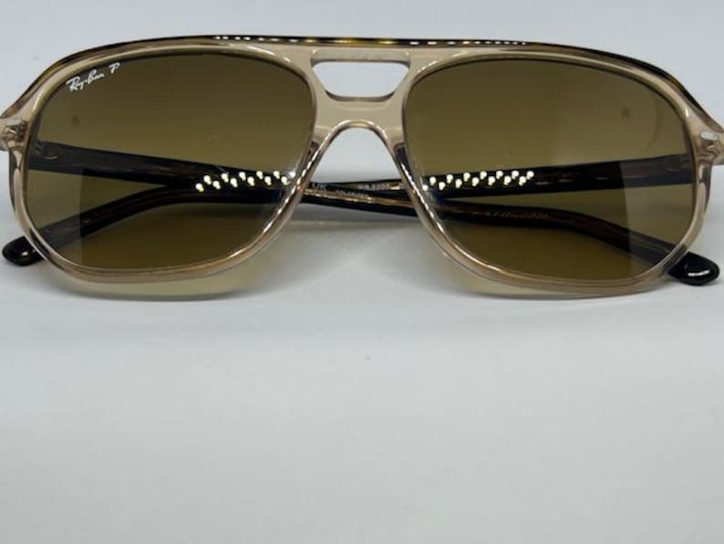May include: Ray-Ban polarised sunglasses featuring a translucent beige plastic frame with a double bridge design and brown tinted lenses. The left lens displays the white Ray-Ban P logo, and the temples are dark brown with a tortoiseshell pattern.