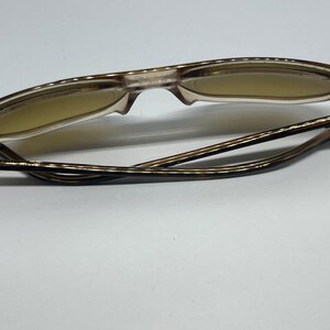 May include: A pair of folded spectacles featuring translucent light brown frames and tinted lenses. The temples exhibit a dark brown tortoiseshell pattern, and the hinges are constructed from silver-toned metal, connecting the frame to the arms.