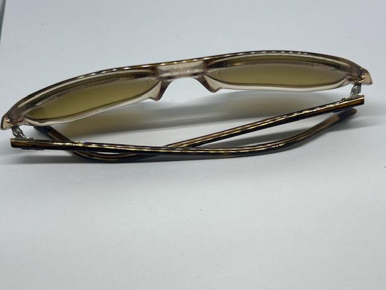 May include: A pair of folded spectacles featuring translucent light brown frames and tinted lenses. The temples exhibit a dark brown tortoiseshell pattern, and the hinges are constructed from silver-toned metal, connecting the frame to the arms.