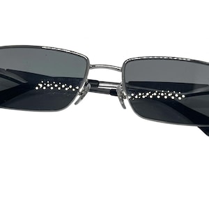 Puede incluir: Gafas de sol rectangulares con una montura met&aacute;lica fina de tono plateado y lentes tintadas en gris oscuro. El dise&ntilde;o incluye almohadillas nasales ajustables y puntas de patillas negras, ofreciendo un aspecto estructurado y limpio.