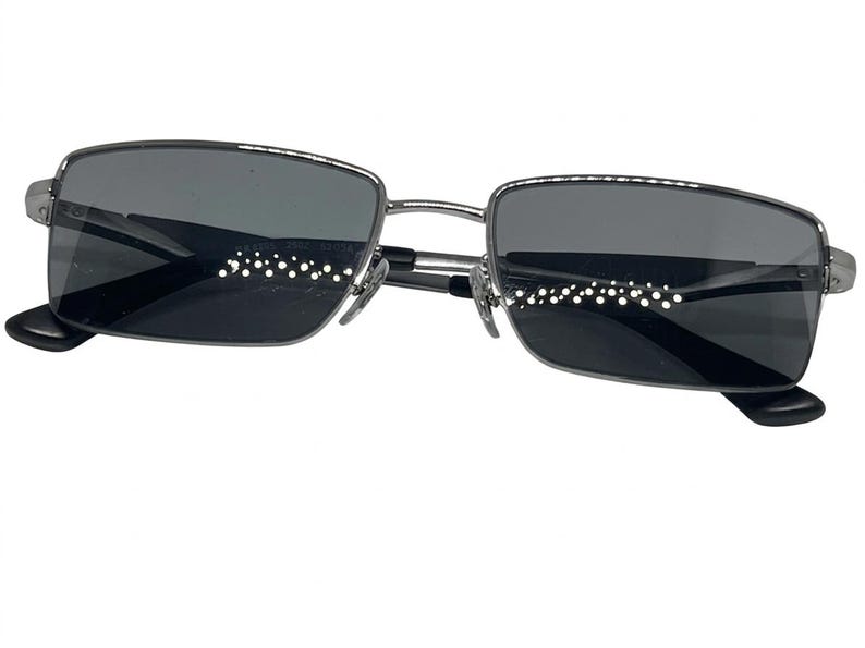 Puede incluir: Gafas de sol rectangulares con una montura met&aacute;lica fina de tono plateado y lentes tintadas en gris oscuro. El dise&ntilde;o incluye almohadillas nasales ajustables y puntas de patillas negras, ofreciendo un aspecto estructurado y limpio.