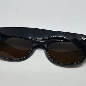 Puede incluir: Gafas de sol Ray-Ban negras con lentes de color marr&oacute;n oscuro. Las monturas tienen un acabado negro brillante con el logotipo plateado de Ray-Ban en ambas patillas. Las gafas est&aacute;n plegadas, mostrando el frente y las patillas.