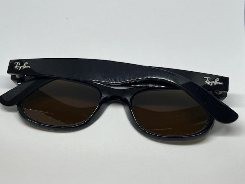 Puede incluir: Gafas de sol Ray-Ban negras con lentes de color marr&oacute;n oscuro. Las monturas tienen un acabado negro brillante con el logotipo plateado de Ray-Ban en ambas patillas. Las gafas est&aacute;n plegadas, mostrando el frente y las patillas.