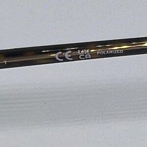 May include: A brown tortoiseshell patterned spectacle temple arm featuring the white printed text Ray-Ban, the CE mark, the UKCA mark, and the word POLARIZED. The material has a glossy finish and a smooth, cylindrical shape with a tapered end.