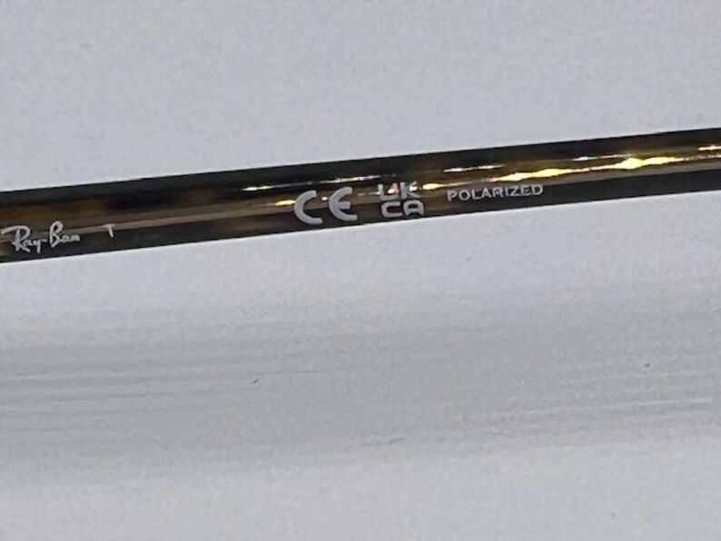 May include: A brown tortoiseshell patterned spectacle temple arm featuring the white printed text Ray-Ban, the CE mark, the UKCA mark, and the word POLARIZED. The material has a glossy finish and a smooth, cylindrical shape with a tapered end.
