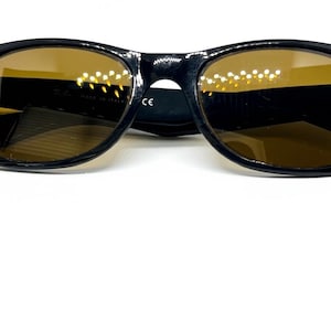 Puede incluir: Gafas de sol negras con monturas ovaladas y lentes tintadas en color &aacute;mbar. Las patillas muestran peque&ntilde;os detalles en tono plateado en las bisagras. El interior de la montura incluye el texto Ray-Ban, Made in Italy y la marca CE visible.