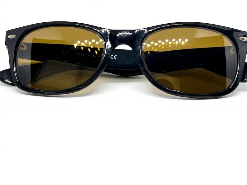 Puede incluir: Gafas de sol negras con monturas ovaladas y lentes tintadas en color &aacute;mbar. Las patillas muestran peque&ntilde;os detalles en tono plateado en las bisagras. El interior de la montura incluye el texto Ray-Ban, Made in Italy y la marca CE visible.
