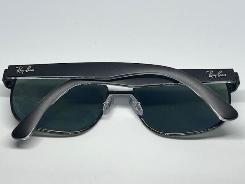 May include: Black Ray-Ban sunglasses featuring dark green lenses and a matte black frame. The temples are folded over the front of the frames, displaying the silver Ray-Ban logo on both the left and right sides of the temple arms.