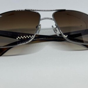 Pode incluir: &Oacute;culos de sol aviador Ray-Ban com uma arma&ccedil;&atilde;o fina em metal prateado gunmetal e design de ponte dupla. As lentes t&ecirc;m um tom castanho degrad&ecirc; e as hastes s&atilde;o feitas de um material castanho escuro com padr&atilde;o de tartaruga e dobradi&ccedil;as.