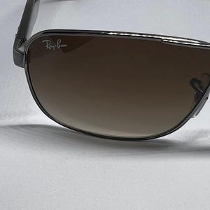 Pode incluir: &Oacute;culos de sol Ray-Ban com arma&ccedil;&atilde;o met&aacute;lica escura e lentes degrad&ecirc; castanhas. A lente esquerda apresenta o log&oacute;tipo branco Ray-Ban no canto superior. As hastes t&ecirc;m um padr&atilde;o de tartaruga escuro e a ponte tem um acabamento met&aacute;lico.